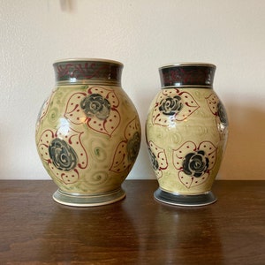 May include: Two ceramic vases with a floral design. The vases are painted with black, green, and red flowers on a cream background.