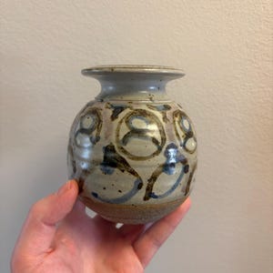May include: A small, round ceramic vase with a wide mouth and a textured, unglazed base. The vase is painted with blue and brown abstract designs on a cream-colored background. The vase is held in a hand.