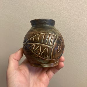 Könnte beinhalten: Eine kleine, braune Keramikvase mit einem stilisierten Fischdesign. Die Vase hat einen runden Körper und eine leicht ausgestellte Öffnung.