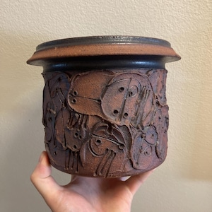 Op de afbeelding: Een handgemaakte, cilindrische keramische pot met een donkerbruine rand en een getextureerd, roodbruin lichaam. De body heeft een reliëf ontwerp van abstracte vormen. De pot wordt in een hand gehouden, wat de grootte en vorm laat zien.