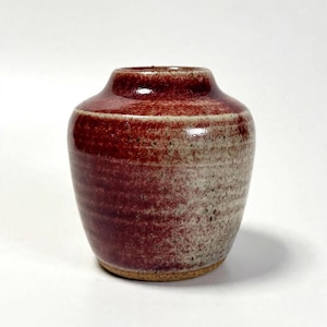 May include: A small, rounded ceramic vase with a glossy finish. The upper portion features a deep, rich red glaze that transitions into a speckled, textured beige and grey base. The vessel has a narrow, circular opening and a thick, sturdy rim.