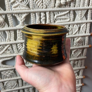 May include: A small, round, ceramic pot with a dark brown and yellow glaze. The pot has a slightly flared rim and a smooth, rounded bottom.