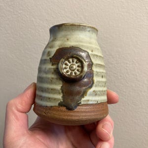 Könnte beinhalten: Eine kleine, weiße Keramikvase mit braunen und weißen Streifen. Die Vase hat ein braunes, kreisförmiges Design mit einer weißen Mitte auf der Vorderseite. Die Vase ist handgefertigt und hat einen rustikalen Look.