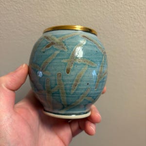 Peut inclure: Petit vase rond en céramique, glaçure bleu clair et bord doré. Le vase est orné d'un motif de coups de pinceau bruns et beiges. Le vase est tenu dans une main.