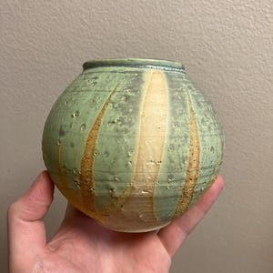 Peut inclure: Un vase rond en céramique fait à la main avec une surface verte tachetée et texturée. Des bandes verticales beiges et marron ornent le vase. Le vase a une petite ouverture et un bord vert foncé.