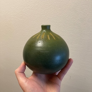 Pode incluir: Um vaso de cerâmica verde escuro artesanal, com corpo redondo e pescoço estreito. O vaso apresenta uma superfície texturizada e um padrão decorativo de linhas amarelas que irradiam do topo. O vaso está na mão de uma pessoa.