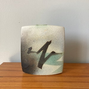 Könnte beinhalten: Eine weiße Keramikvase mit einem abstrakten Design in Schwarz und Grün. Die Vase hat eine rechteckige Form mit abgerundeten Kanten.