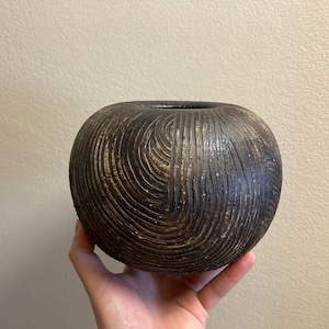 May include: A dark brown, spherical ceramic vase with a textured, carved surface. The vase has a wide opening at the top and is held in a hand. The carved pattern creates a swirling, organic design.