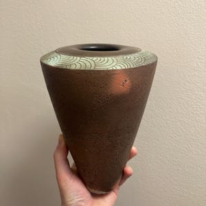 May include: A brown ceramic vase with a green and white geometric design around the top rim. The vase is cone-shaped and has a textured surface.