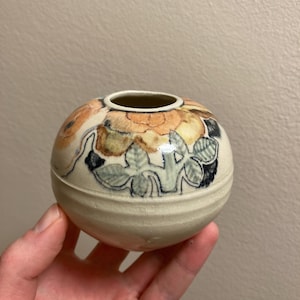 Pode incluir: Um pequeno vaso redondo de cerâmica branca com um desenho floral em tons de laranja, amarelo, verde e preto.