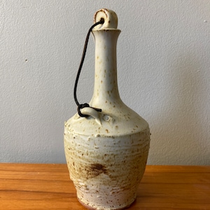 Op de afbeelding: Een witte keramische fles met een bruine glazuur en een metalen handvat. De fles heeft een rond lichaam en een lange, dunne hals.