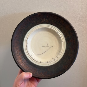 May include: A handcrafted ceramic plate with a dark brown rim and a cream-coloured centre. The centre has a simple black line drawing and markings. The plate is held by a hand.
