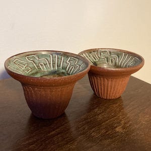 May include: Two terracotta ceramic bowls with a green glaze and a white design. The bowls are sitting on a wooden surface.