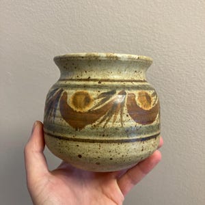 Puede incluir: Un jarrón pequeño, redondo, de cerámica con un esmalte marrón moteado. El jarrón tiene un diseño marrón, naranja y verde alrededor del medio.