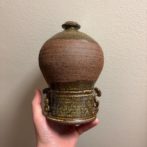 May include: A handcrafted ceramic jar with a lid. The jar has a bulbous body with a textured, brown band around the middle. The base and lid are a darker, speckled green-brown. Two decorative handles are on the base.