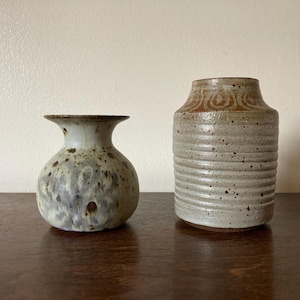 Könnte beinhalten: Zwei Keramikvasen, eine mit einem gesprenkelten braunen und weißen Überzug und die andere mit einem weißen Überzug und braunen Linien. Beide Vasen stehen auf einer dunkelbraunen Holzoberfläche.