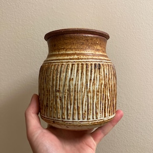 Könnte beinhalten: Eine braune und weiße Keramikdose mit Deckel. Die Dose hat ein strukturiertes, gestreiftes Design.