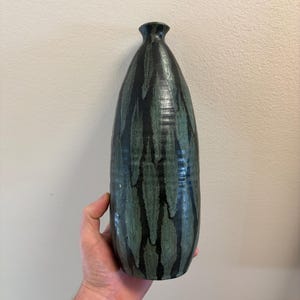 May include: A tall, slender ceramic vase with a narrow neck and a flared rim. The surface features a dark black base glaze with vertical, streaky patterns of muted teal and sage green, creating a textured, layered appearance across the body.