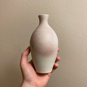 May include: A small, white ceramic vase with a subtle pink ombre effect. The vase has a rounded, bulbous shape and a narrow neck.