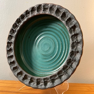 Peut inclure: Un bol rond en céramique avec un glaçage turquoise et un bord noir texturé. Le bol est posé sur un support en acrylique transparent.