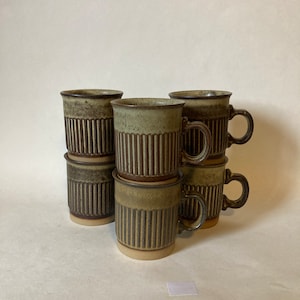 Op de afbeelding: Set van zes bruine en groene gestreepte keramische mokken met handvatten. De mokken zijn in een piramidevorm gestapeld.