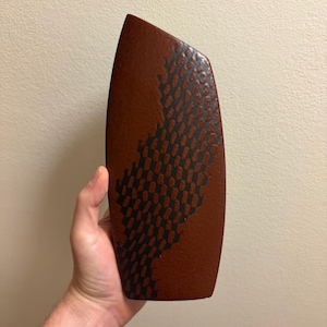 May include: A tall, rectangular ceramic vase with a textured, matte rust-brown finish. The front surface features a prominent, vertical, dark charcoal-coloured pattern consisting of repeating, slightly irregular, elongated oval shapes.