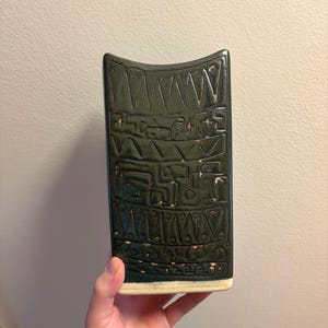 Könnte beinhalten: Dunkelgrüne Keramikvase mit rechteckiger Form und gewölbter Oberseite. Die Vase hat ein geprägtes geometrisches Muster in einem helleren Grünton. Der Sockel ist cremefarben.