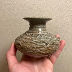 May include: A small, handcrafted ceramic vase with a textured, wave-like pattern on the body. The vase has a wide, rounded base and a narrow, flared opening. The colors are earthy, with shades of brown, grey, and tan.