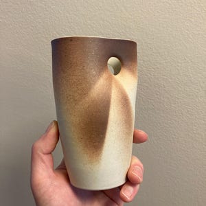 May include: A white ceramic vase with a brown glaze and a hole in the side. The vase has an irregular shape and a textured surface.