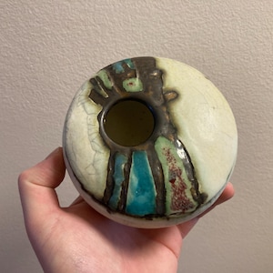 Op de afbeelding: Een ronde keramische vaas met een centrale opening. De vaas heeft een crèmekleurige buitenkant met bruine, blauwgroene en rode accenten. Het ontwerp omvat een gestructureerd, gekraakt effect, wat bijdraagt aan de unieke esthetiek. Dit decoratieve stuk is ideaal voor interieurdecoratie.