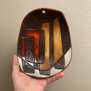 Peut inclure: Une suspension murale en céramique avec un motif géométrique dans les tons de brun, orange et blanc. La suspension a une surface texturée et un trou en haut pour la suspendre.