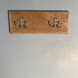 Peut inclure: Un porte-manteau en bois avec deux crochets en métal argenté. Le support est rectangulaire avec une finition en grain de bois brun clair. Les crochets sont fixés sur le panneau de bois et sont conçus pour suspendre des manteaux, des chapeaux ou d'autres articles. Il est monté sur un mur bleu clair.