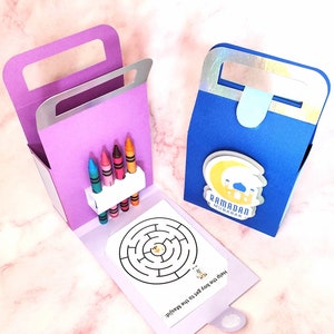 May include: Two colorful cardboard boxes with a Ramadan Mubarak design. The box on the left has a built-in holder for crayons and a maze activity. The box on the right is blue with a crescent moon and star design.
