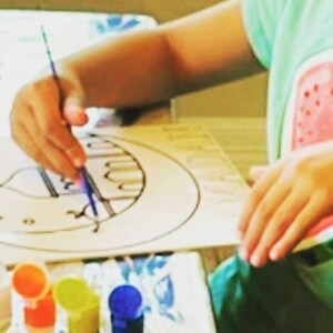 May include: A child uses a paintbrush to color a black and white drawing of a mosque on a white paper. The drawing is surrounded by small stars and circles. The child is wearing a green shirt with a watermelon design. There are several small bottles of paint in the foreground.