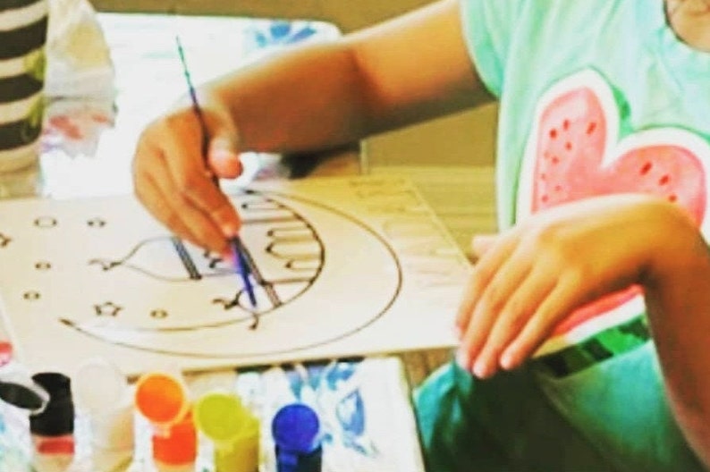 May include: A child uses a paintbrush to color a black and white drawing of a mosque on a white paper. The drawing is surrounded by small stars and circles. The child is wearing a green shirt with a watermelon design. There are several small bottles of paint in the foreground.