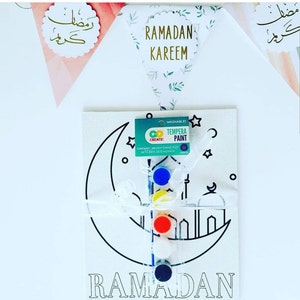 May include: A white canvas with a black outline of a crescent moon and a mosque. The canvas is labeled "RAMADAN". A set of washable tempera paint pots with a brush is included. The paint pots are red, blue, yellow, white, and black.