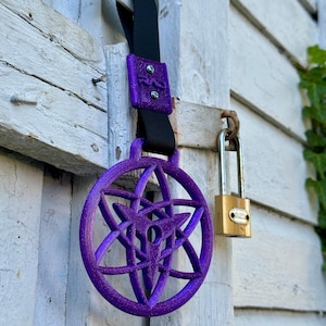 Op de afbeelding: Een paars, stervormig decoratief item met een zwarte band en een paarse sluiting. Het item hangt aan een witte houten deur. Een gouden hangslot is ook zichtbaar.