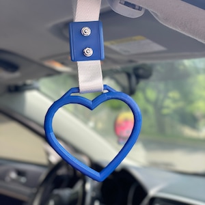 May include: A blue heart-shaped car handle with a white strap and a blue plastic clip. The handle is attached to the ceiling of a car.