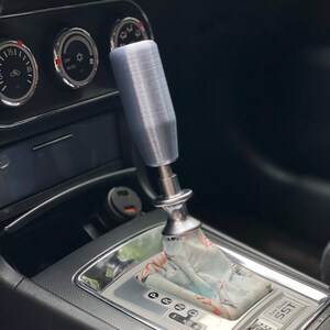 Puede incluir: Primer plano de la palanca de cambios de un coche con una perilla de cambio plateada y una funda de palanca de cambios con estampado. La palanca de cambios tiene un ribete plateado y el texto "Twin Clutch SST" es visible.