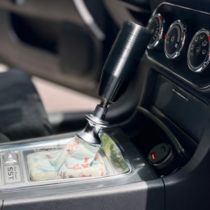 Puede incluir: Primer plano de la palanca de cambios de un coche con una perilla de cambio negra y una funda de palanca de cambios con estampado. La palanca de cambios est&aacute; etiquetada como "SST" y "Top Clutch".
