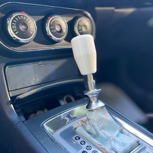Puede incluir: Una perilla de palanca de cambios blanca con una base plateada, instalada en la consola central de un autom&oacute;vil. La palanca de cambios tiene una funda con estampado y un borde plateado alrededor del selector de marchas.