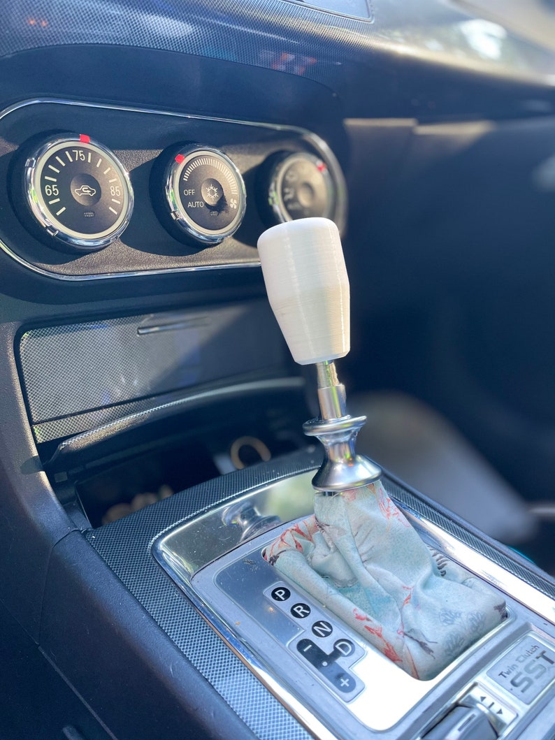 Puede incluir: Una perilla de palanca de cambios blanca con una base plateada, instalada en la consola central de un autom&oacute;vil. La palanca de cambios tiene una funda con estampado y un borde plateado alrededor del selector de marchas.