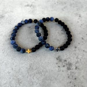 May include: Two stretch bracelets made of round beads. One bracelet features blue and grey mottled beads with a gold-toned cross charm. The second bracelet consists of black textured beads and blue mottled beads with a silver-toned cross charm.