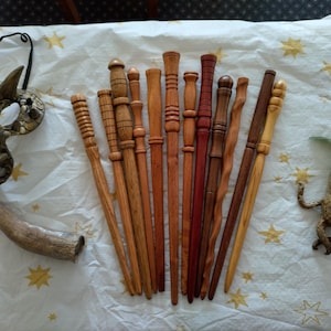 Puede incluir: Una colección de varitas de madera en varios tonos marrones, junto con una máscara tallada, un cuerno y una pequeña figura de mandrágora. Las varitas varían en diseño, algunas con tallas intrincadas y otras con un acabado liso.