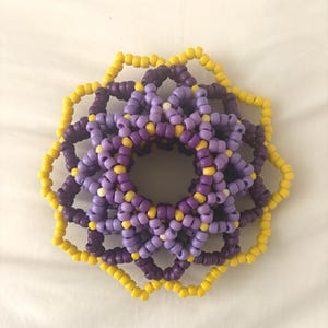 May include: A beaded hair accessory in a floral design. The accessory features a layered pattern of yellow, purple, and lavender beads. The outer layer is yellow, followed by a layer of dark purple, and the center is lavender.