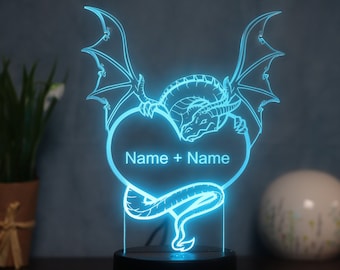 Op de afbeelding: Een blauwe lichtgevende draak die een hart vasthoudt met de tekst "Name + Name" in het midden. De draak houdt het hart vast met zijn klauwen en staart. Het licht is aan en verlicht de draak en het hart.