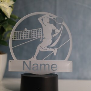 Op de afbeelding: Witte silhouet van een tennisspeler met een racket en een bal, een tennisnet en het woord "Name" op een witte basis. De basis staat op een zwarte ronde basis.