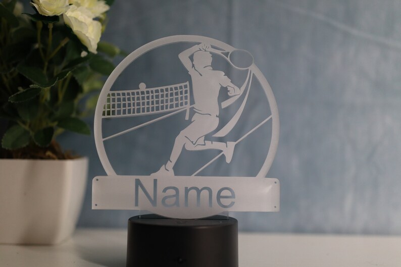 Op de afbeelding: Witte silhouet van een tennisspeler met een racket en een bal, een tennisnet en het woord "Name" op een witte basis. De basis staat op een zwarte ronde basis.