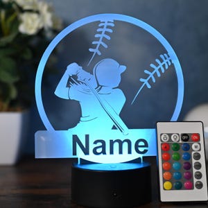 Peut inclure: Une veilleuse personnalisée bleue et blanche avec une silhouette de joueur de baseball et un graphique de baseball. La lumière est dotée d'une télécommande avec plusieurs options de couleur. Le nom "Name" est affiché sur la base de la lumière.