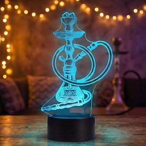 May include: A 3D illusion LED lamp featuring a glowing cyan line-art design of a hookah. The acrylic panel displays a detailed silhouette of the water pipe, hose, and bowl, mounted on a circular black base with a power button on the front.